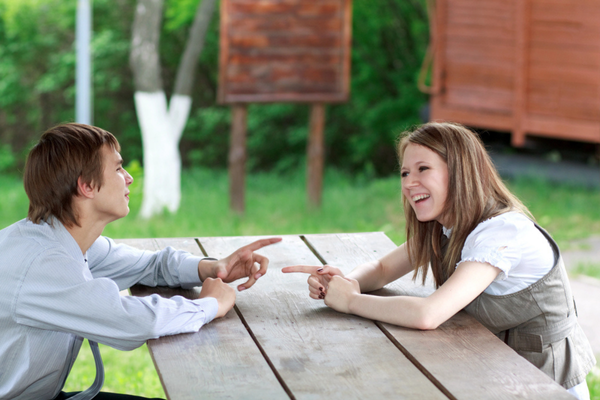Two teenagers sit outdoors at a picnic table, laughing and pointing while having a fun conversation
