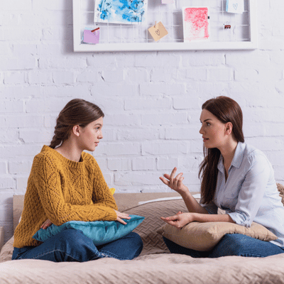 Mother and teenage daughter sit on bed, exchanging concerns and advice during calm parenting conversation