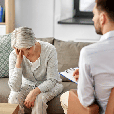 Concerned woman holds head during therapy appointment while counselor listens and takes notes empathetically today