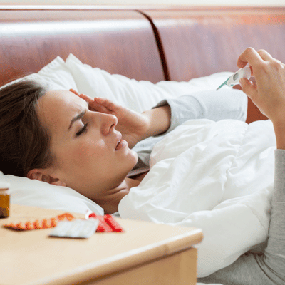 Sick woman checks thermometer in bed, considering hoarse voice causes and nearby medications for relief