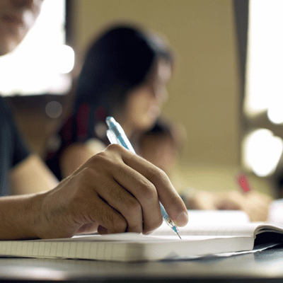 Close-up hand writes with pen in notebook during lecture while classmates study in background attentively