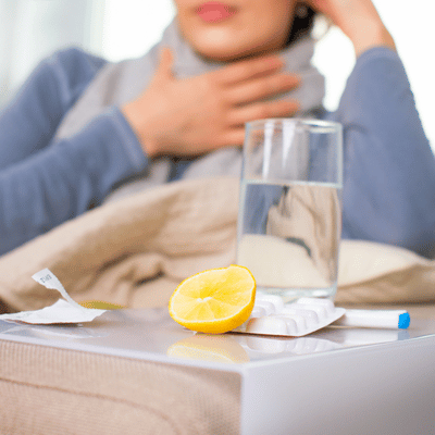 Glass of water, lemon, and tablets on table; ill woman clutches throat in background today