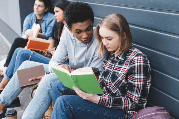 Students sitting against wall share green textbook, studying together outside school building between classes today