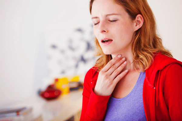 Woman touches throat, wincing, showing hoarseness symptoms in a home setting portrait during seasonal illness