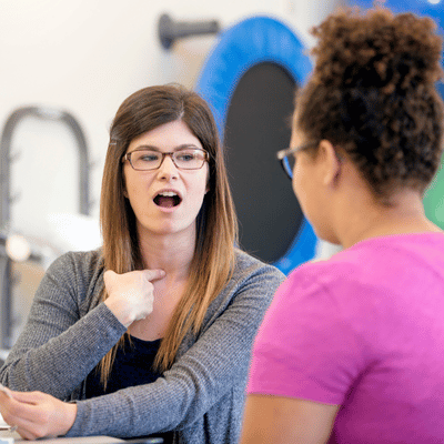 Speech therapist guides adult woman pointing to throat during private speech therapy articulation exercise session