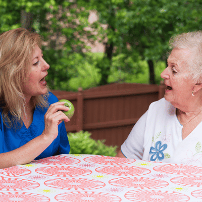Therapist practices mouth movements with senior woman outdoors during private speech therapy using visual cue