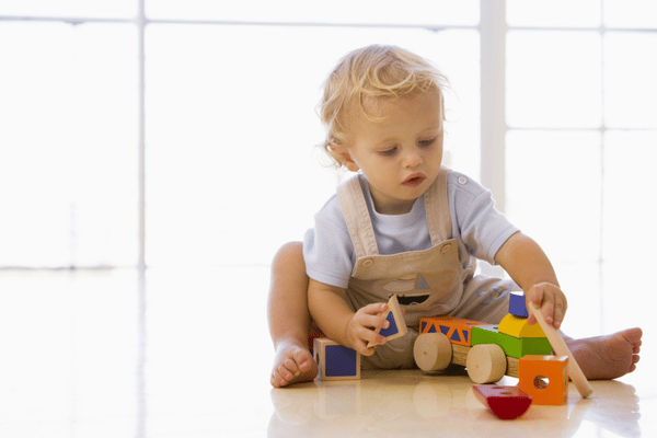 Toddler sits on floor stacking wooden toys, building skills before speech therapist for 2-year-old support